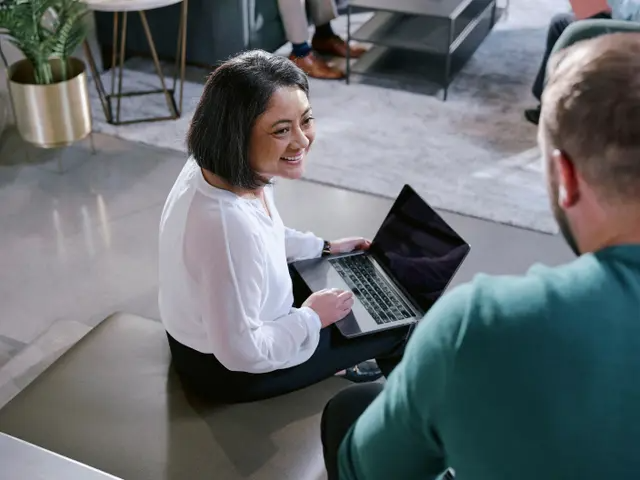 A woman sitting down with a laptop on her lap talking to a man standing near her.
